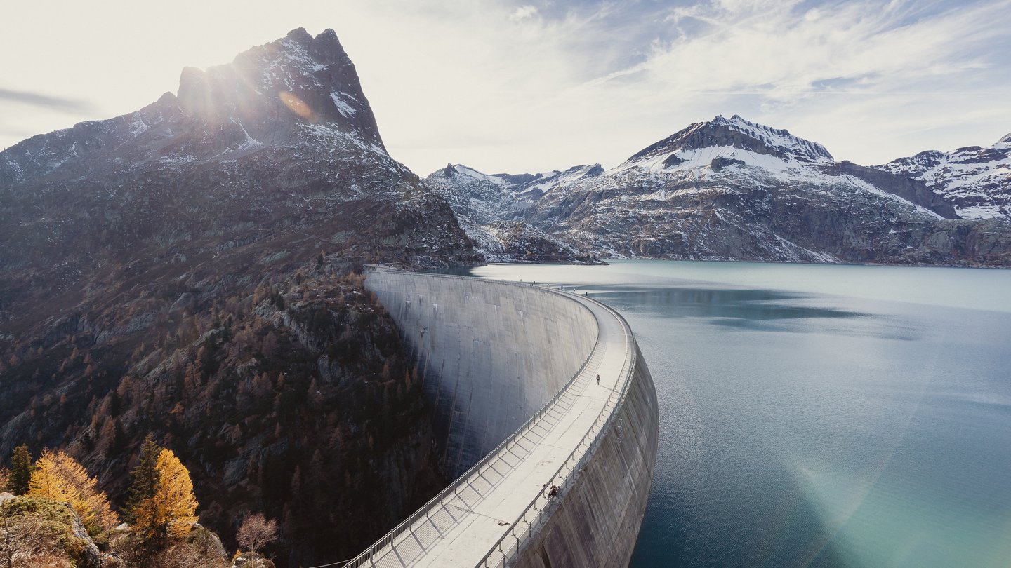 A large curved dam stretches across an alpine valley, holding back a turquoise reservoir surrounded by snow-capped mountains. The sun shines from behind a rugged mountain peak in the background.