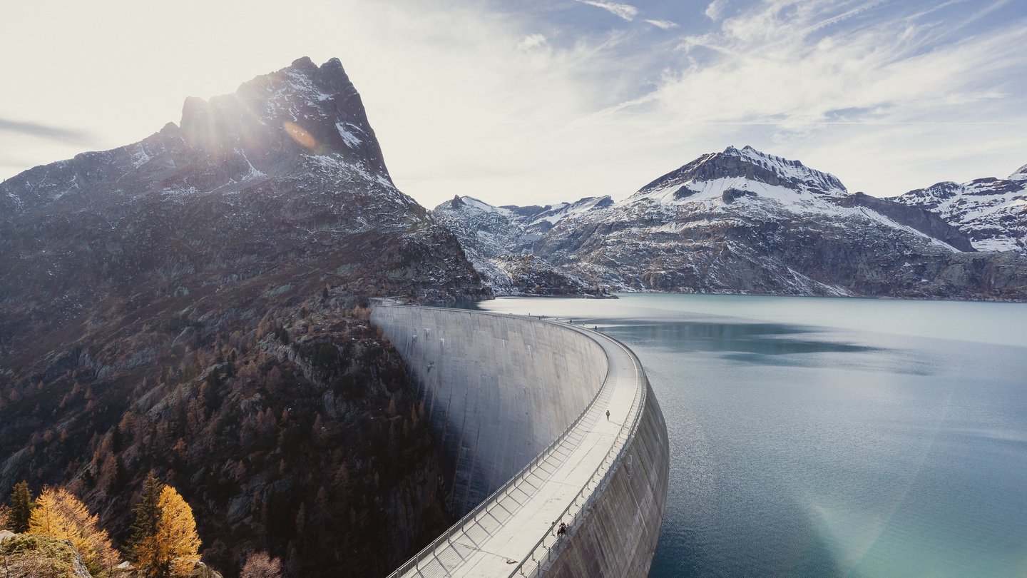 A large curved dam stretches across an alpine valley, holding back a turquoise reservoir surrounded by snow-capped mountains. The sun shines from behind a rugged mountain peak in the background.
