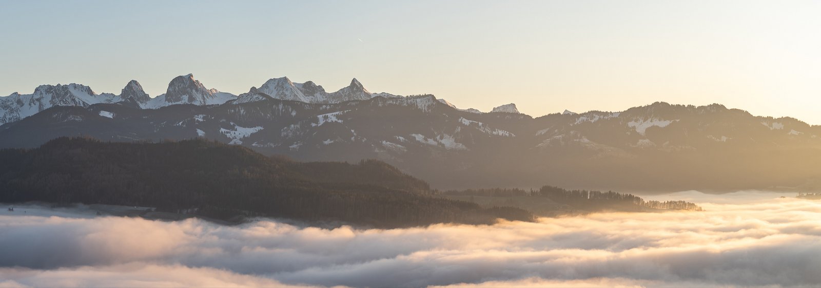 Schneebedeckte Bergkette über einem Nebelmeer, beleuchtet von warmem Sonnenlicht.