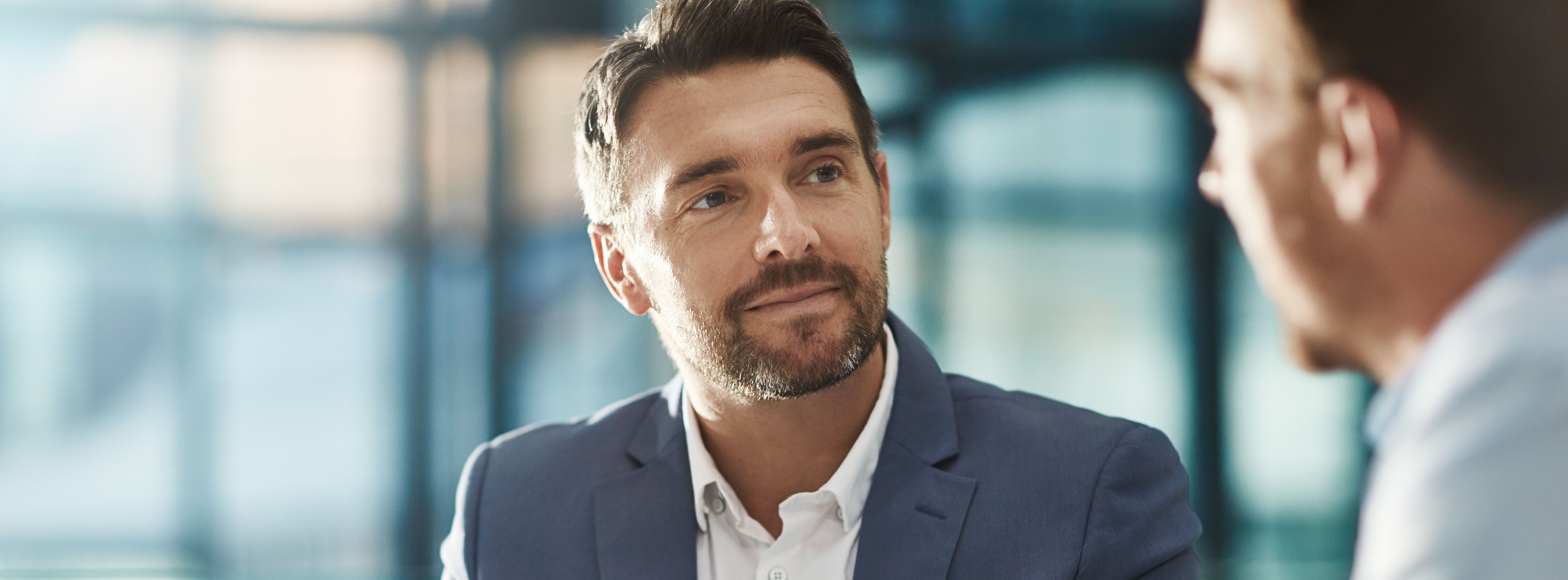 Ein Mann im Anzug und weissem Hemd schaut im Rahmen eines Beratungsgespräches rüber zu einem anderen Mann und hört aufmerksam zu